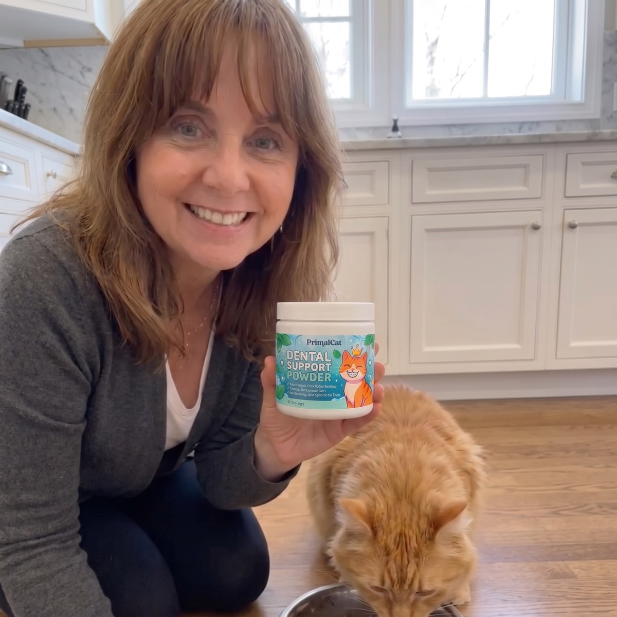 A woman crouching in her kitchen holding a tub of PrimalCat Dental Support Powder while her orange cat eats from a bowl beside her.